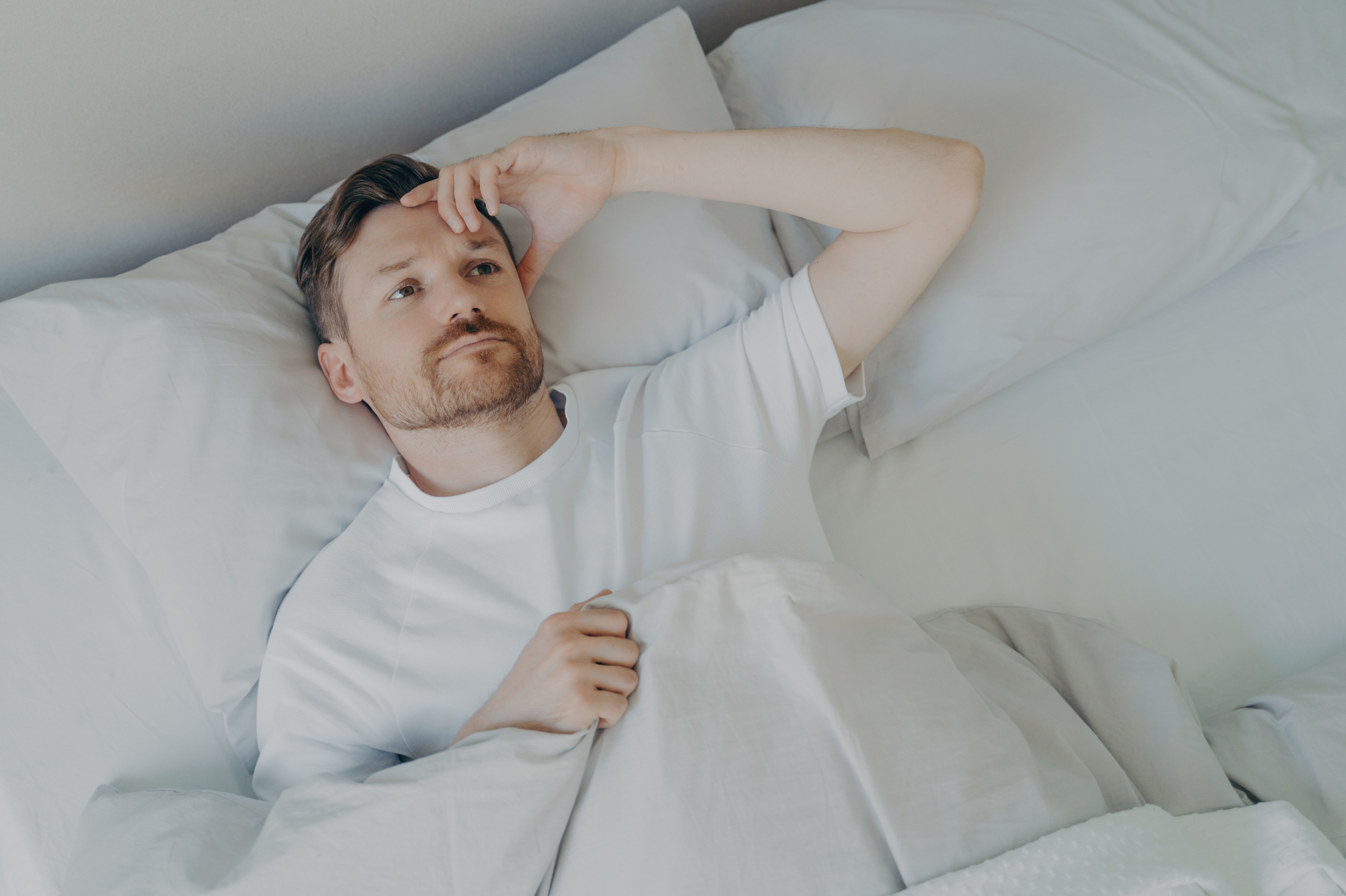 A bearded man lies in bed with insomnia, worrying about sleep’s effect on athletic performance.