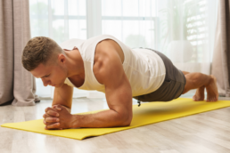 A man in a white tank top holds a static plank on a yellow yoga mat to improve his core strength for cycling.