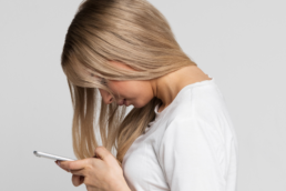 A blonde woman suffering from text neck stands with poor posture looking down at her cell phone.