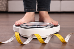 Two feet stand on a scale with a tape measure curled on the ground nearby, representing BMI vs. body composition.