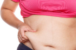 A woman in a pink shirt pinches some excess body fat around her waist, representing a discussion on the types of body fat.
