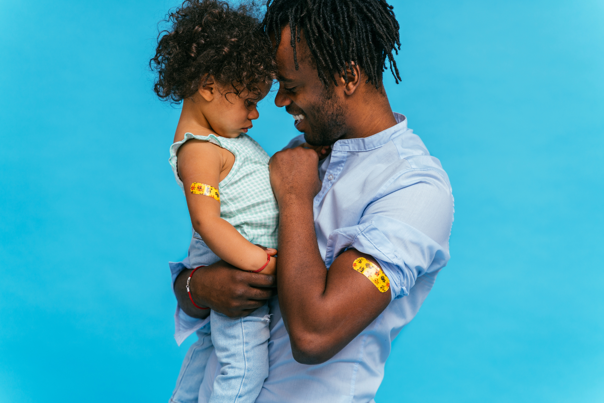 A father and daughter stand against a blue background with yellow band-aids, after receiving vaccines.