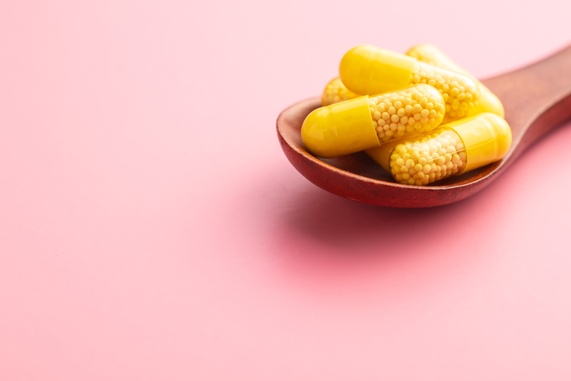 Yellow vitamins for anti aging sit on a wooden spoon in front of a light pink background.