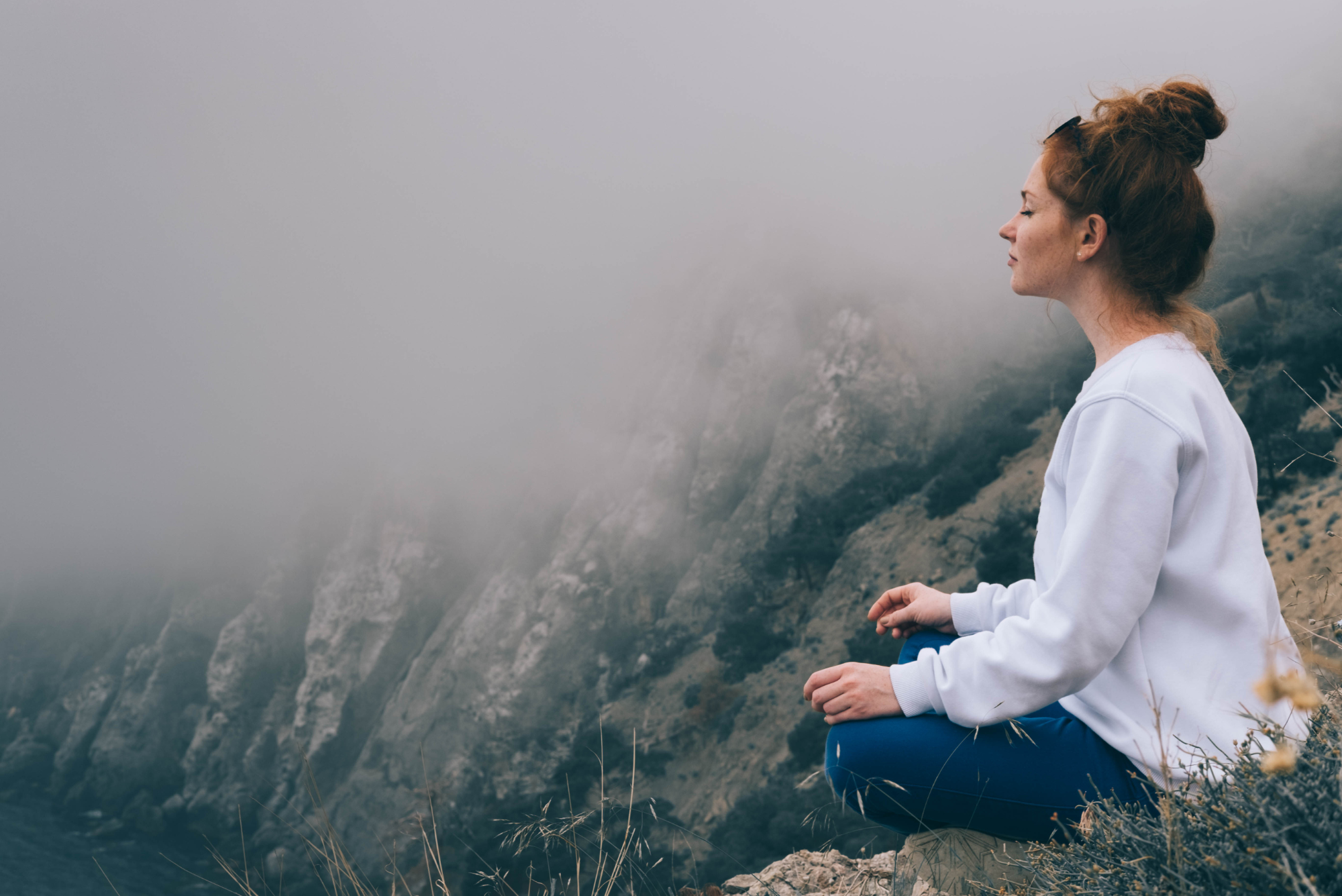 A woman in a white sweatshirt sits meditating atop a hill on a misty day, following Banner Peak’s 12-step meditation guide.
