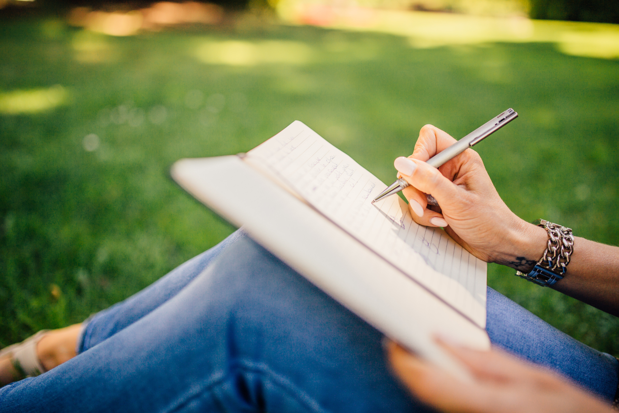 A woman sits in a park on the green grass, journaling to cope with stress and anxiety.