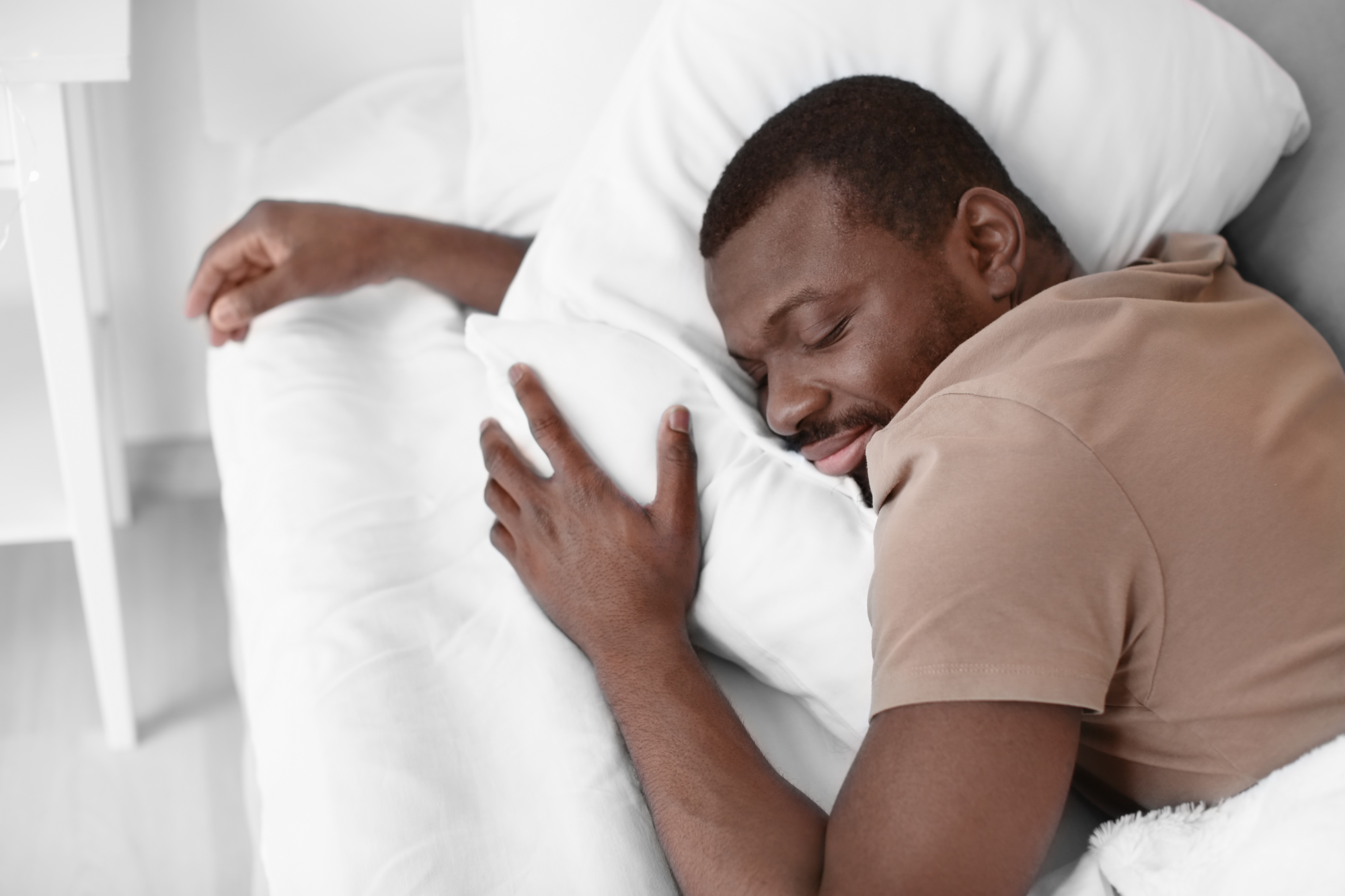 A man smiles while sleeping peacefully on a bed covered in plush white pillows.
