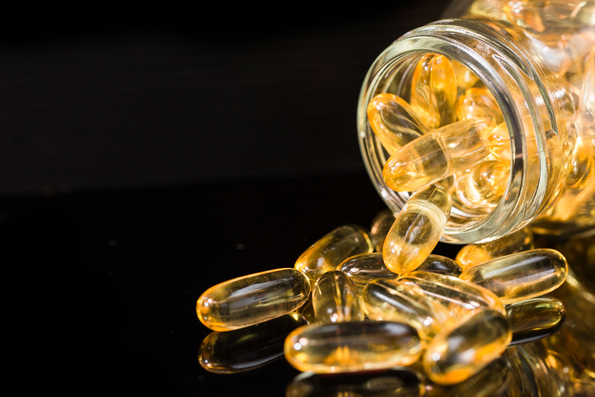 A handful of fish oil capsules spill out of a clear glass pill bottle that is tipped over on a black tabletop.