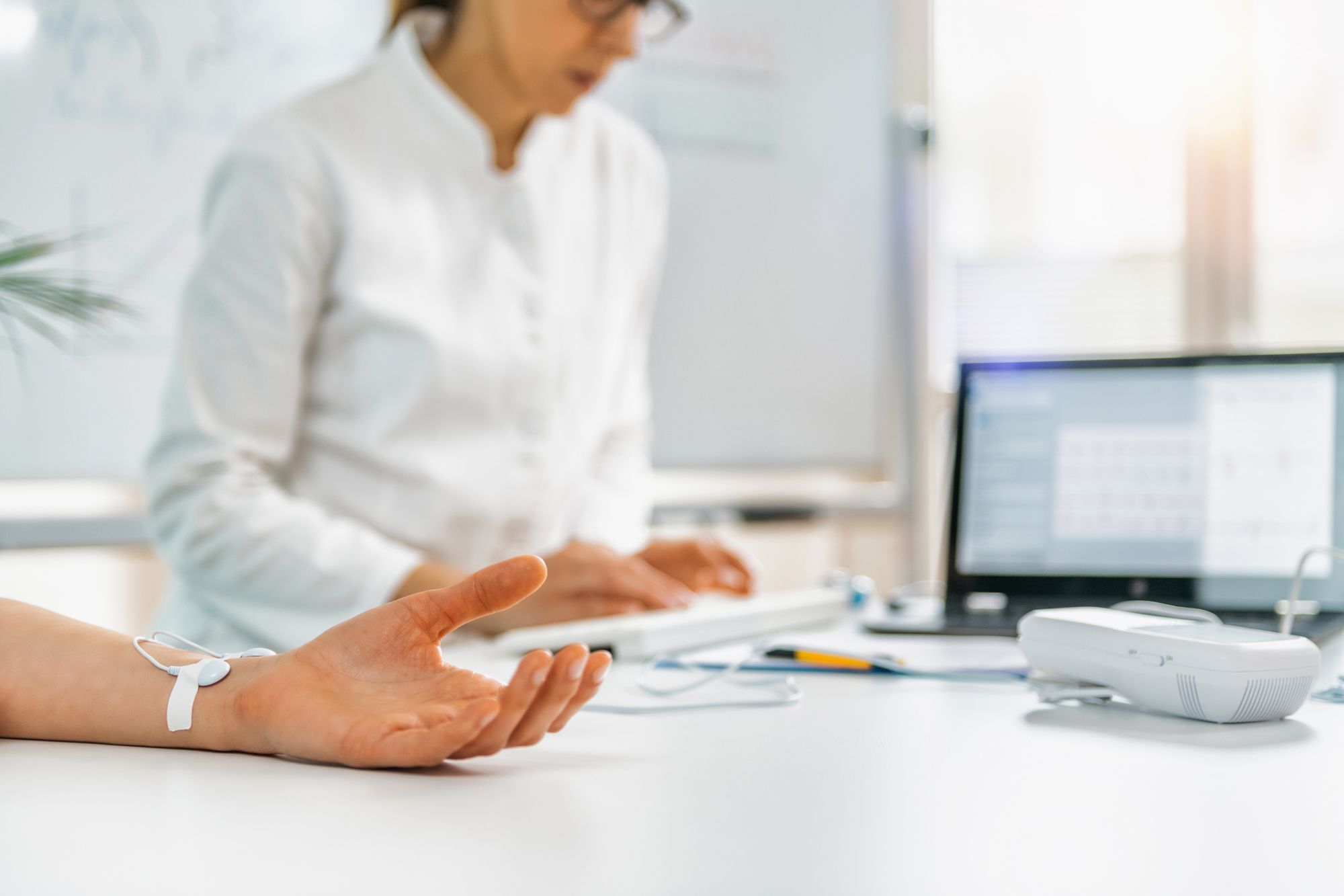 A doctor is monitoring a patient’s heart rate with a sensor attached to their hand while recording data on a computer.