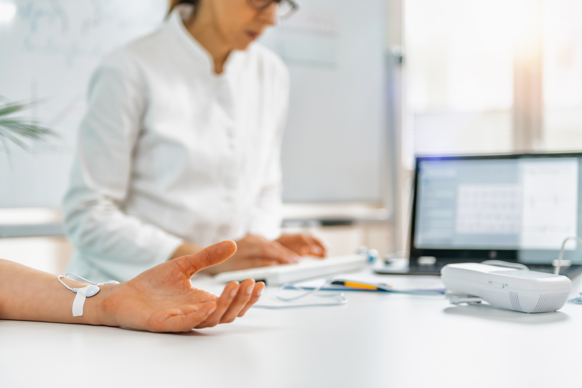 A doctor is monitoring a patient’s heart rate with a sensor attached to their hand while recording data on a computer.