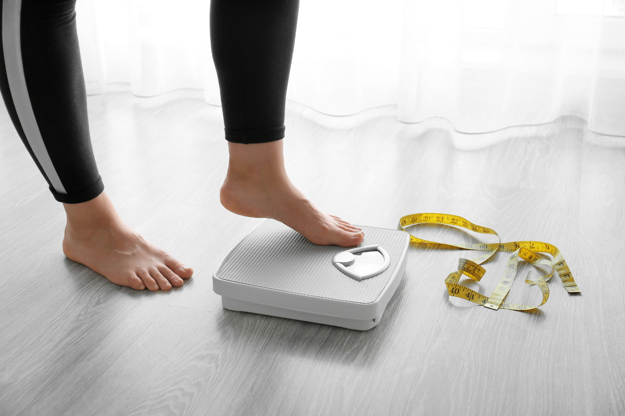 A person stepping onto a scale with a measuring tape on the floor, indicating a focus on weight loss.