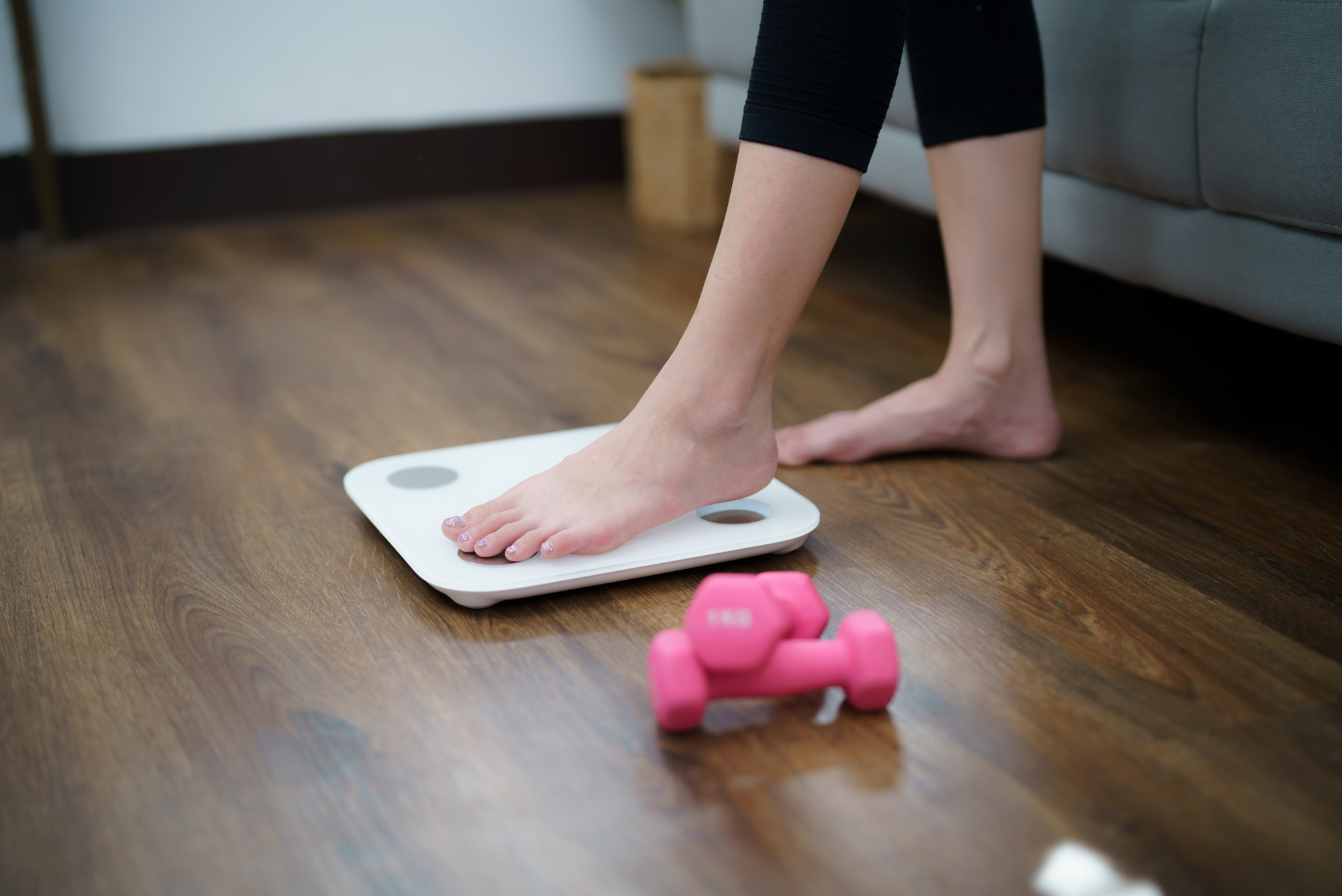 Bare feet on white smart scale with pink dumbbells on wooden floor, showing weight management and strength training elements.