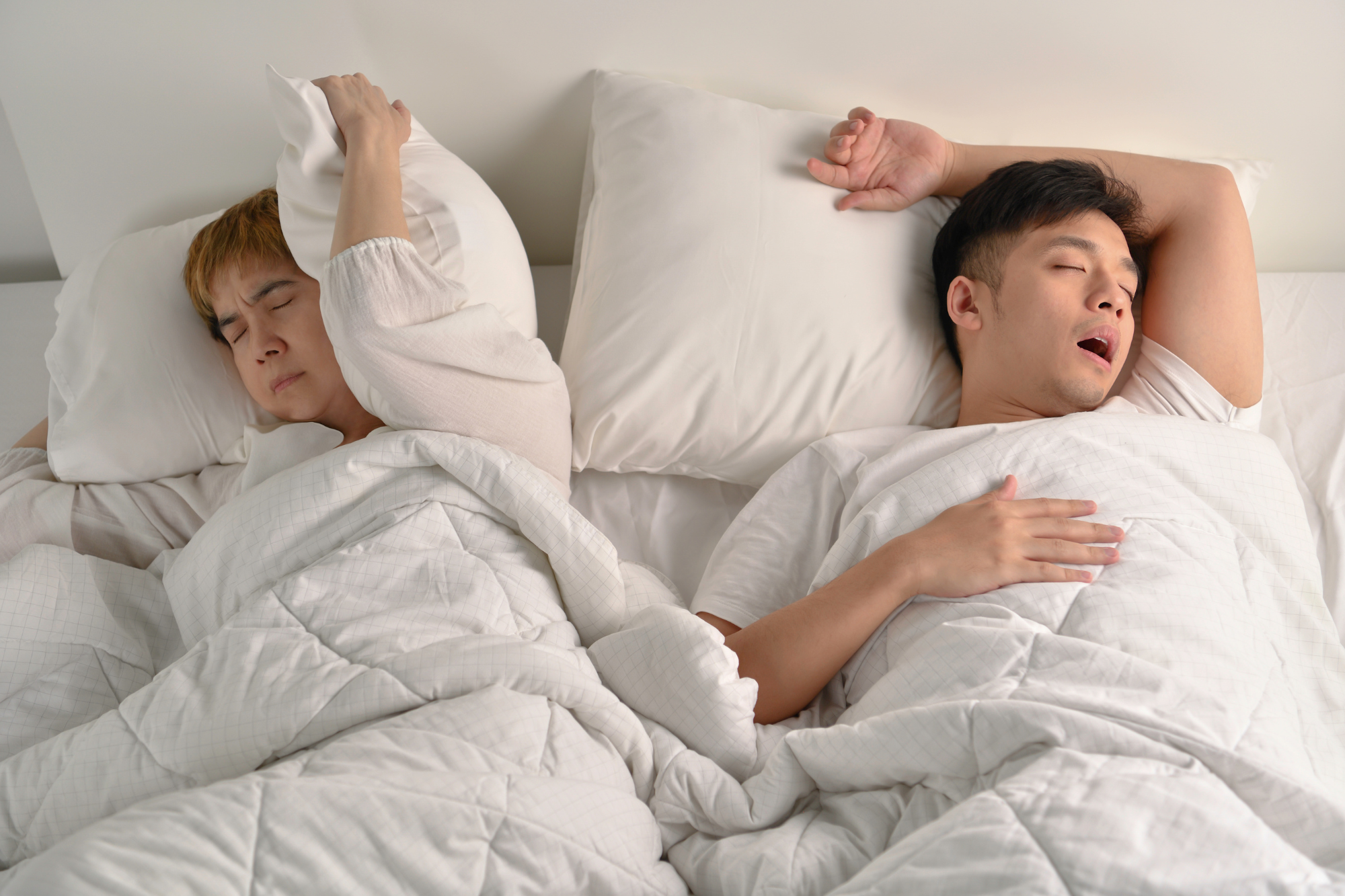 Two people sleeping in bed, one covering ears with pillow while other snores.