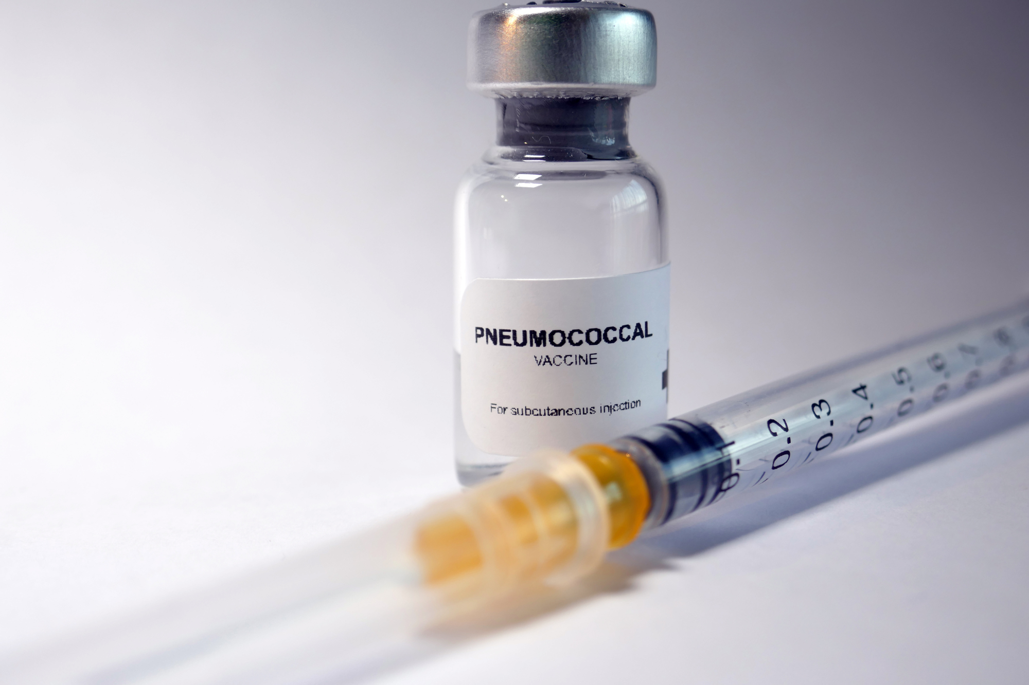 Close-up of a pneumococcal vaccine labeled for subcutaneous injection, alongside a medical syringe on a white background.