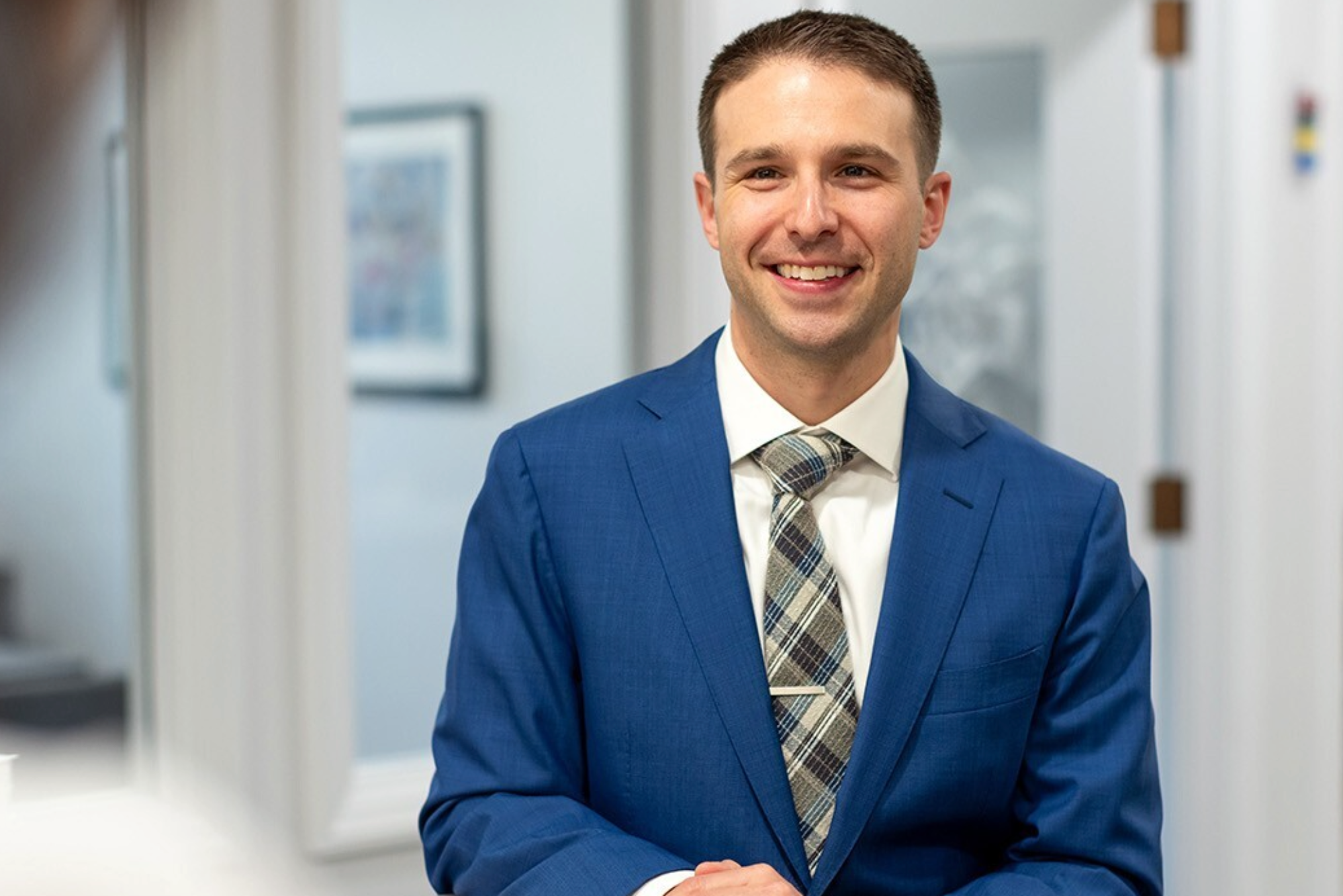 Smiling man in a blue suit and plaid tie standing in a professional office hallway.