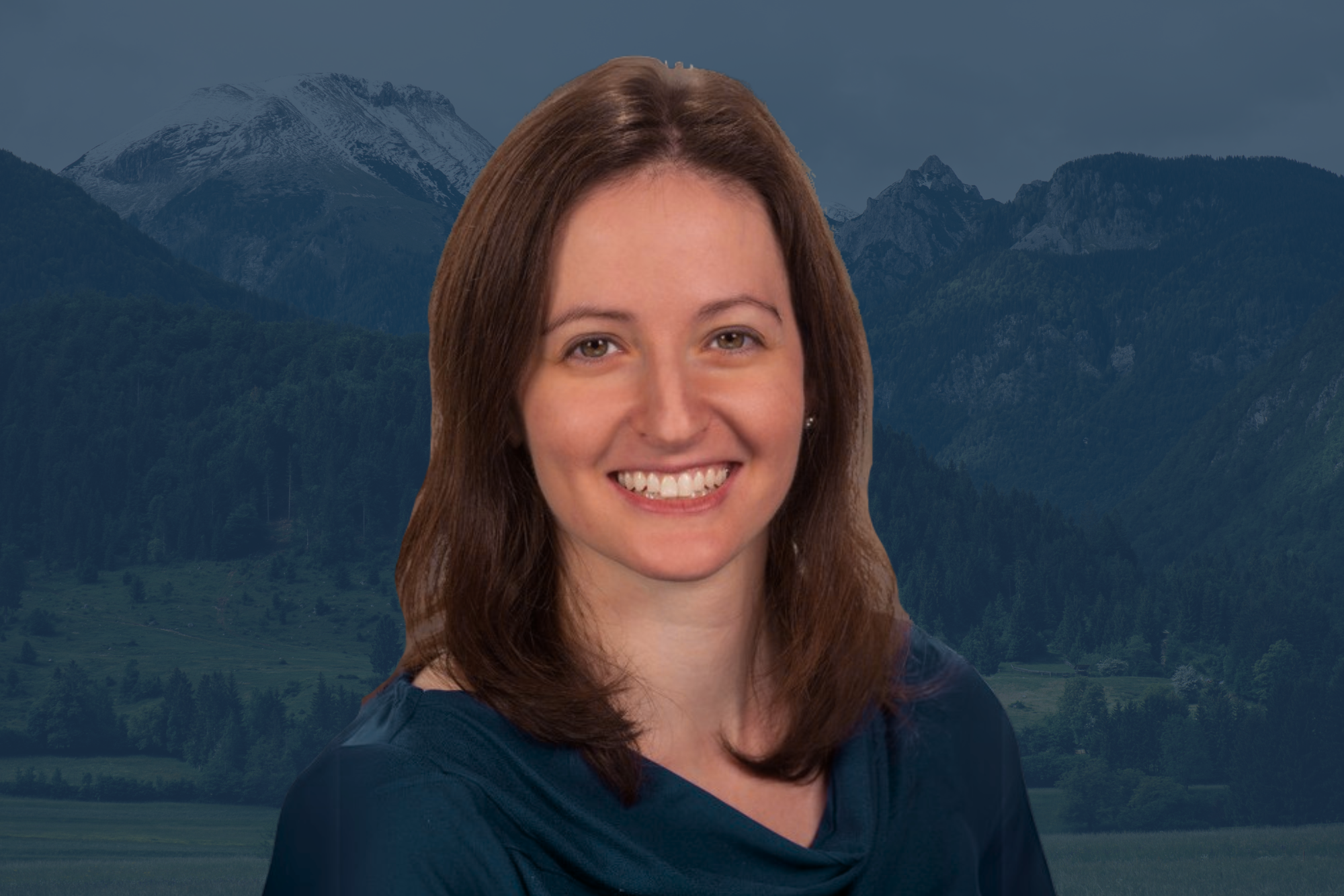 A woman with shoulder-length brown hair smiling, wearing a dark blue top, with forested mountains in the background.