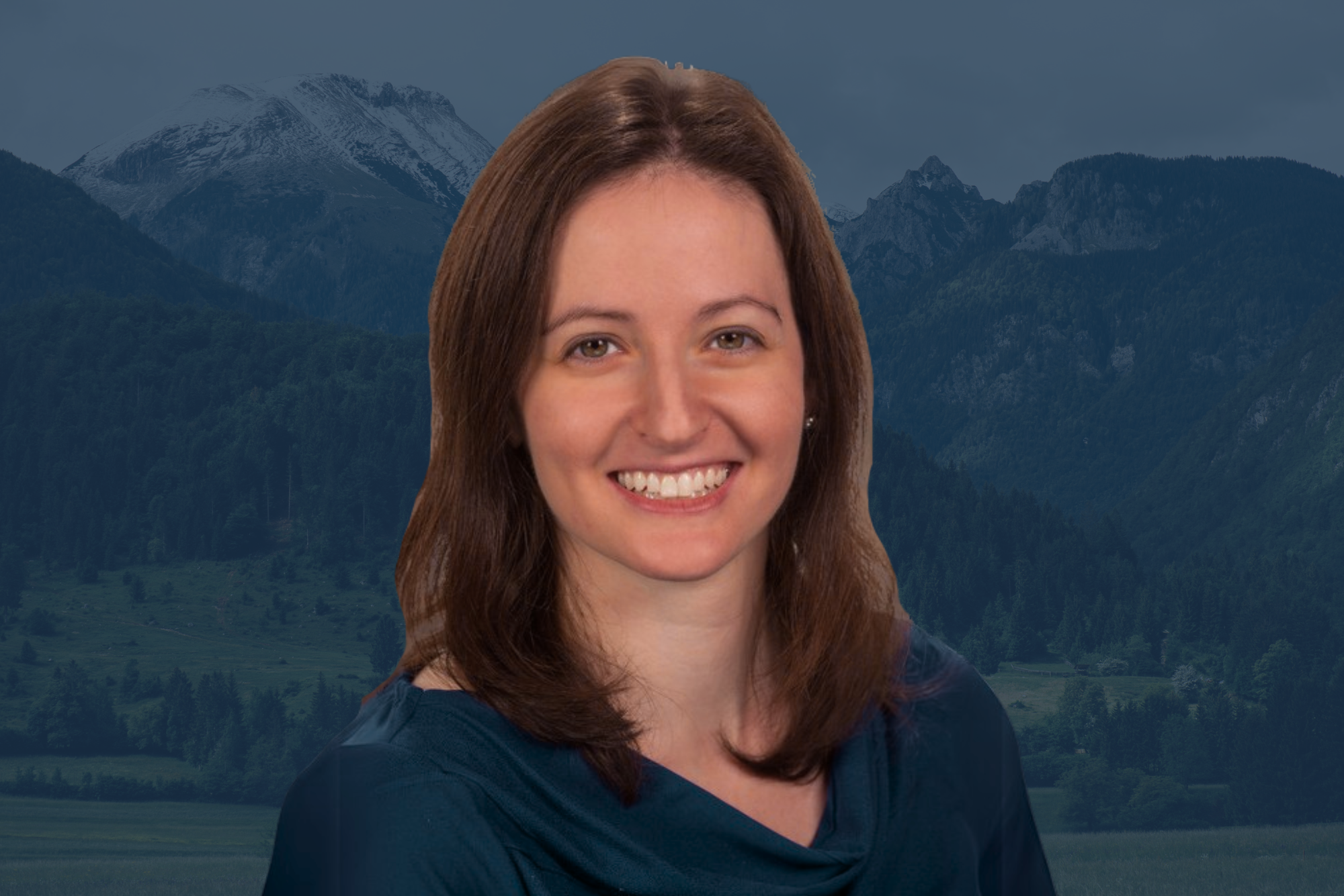 A woman with shoulder-length brown hair smiling, wearing a dark blue top, with forested mountains in the background.