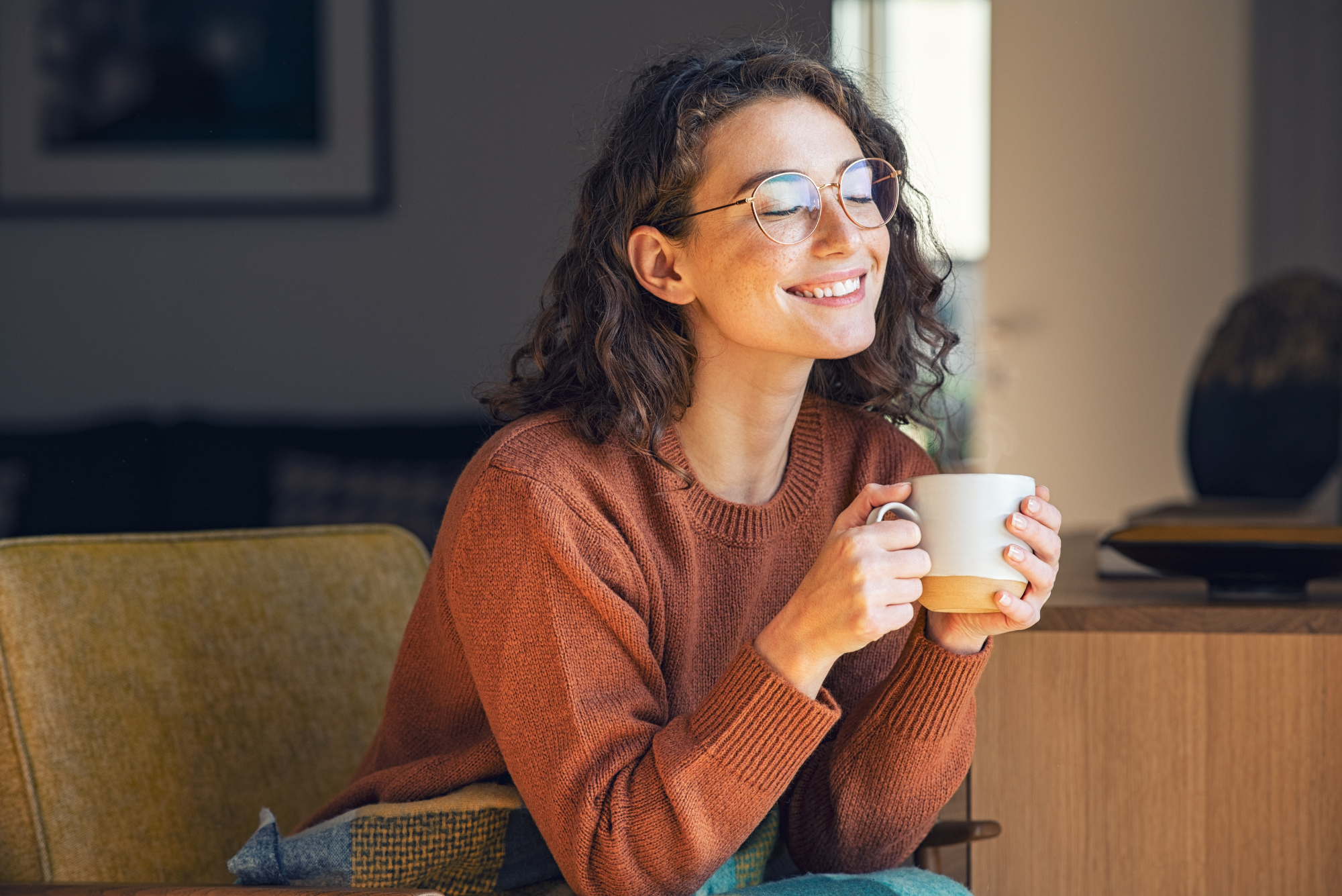 Smiling woman with curly hair and glasses enjoying a warm drink, sitting relaxed indoors.