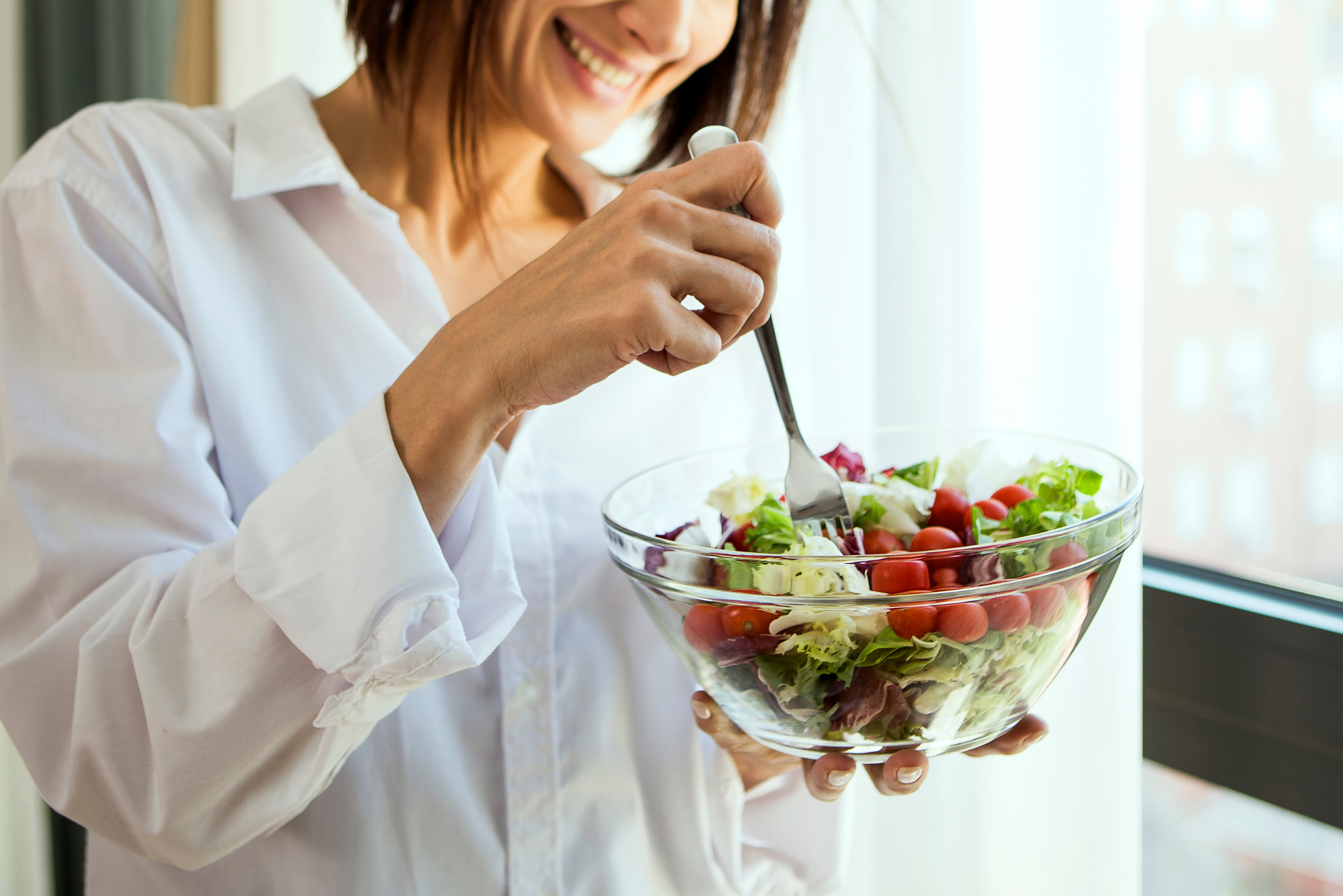 Smiling woman eats colorful, fresh vegetable salad from glass bowl.