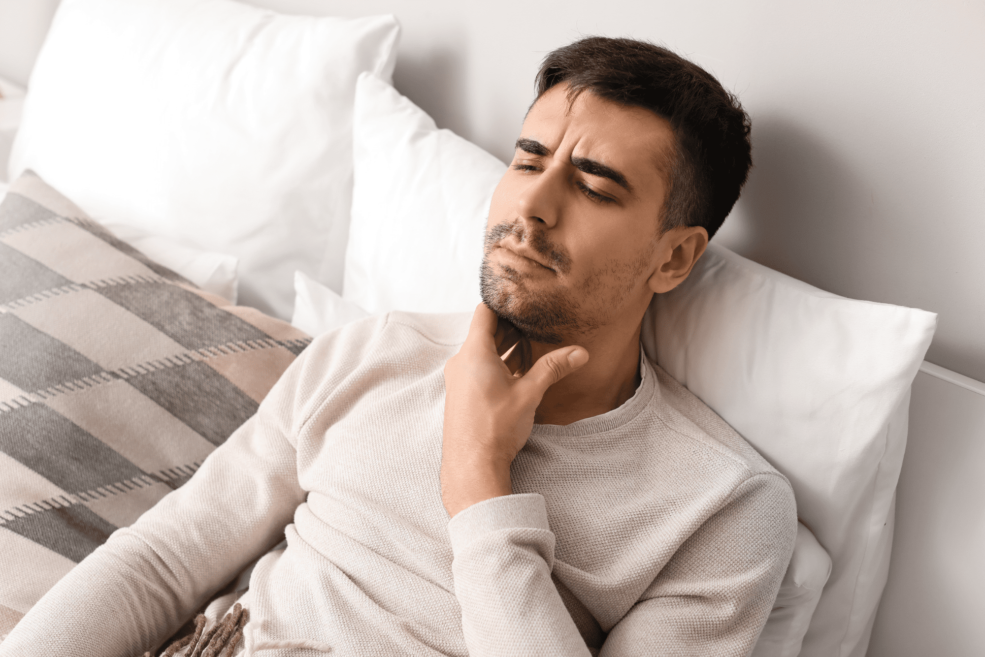 Close-up of man lying in bed with pained expression, suggesting morning throat discomfort.