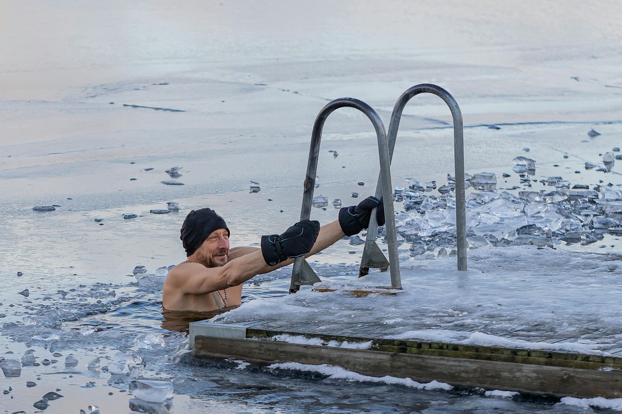 A man in a beanie hat and swim trunks immersed in frigid water up to his chest.