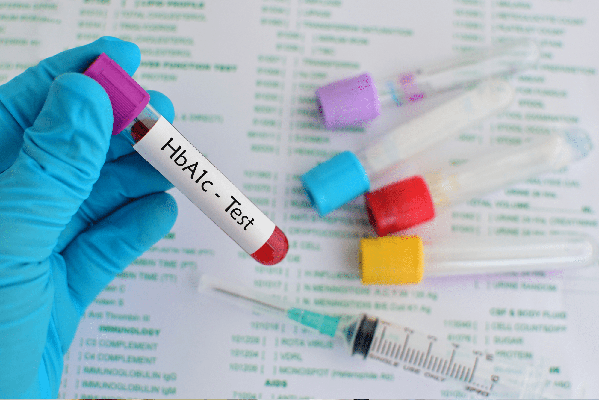 Gloved hand holds blood vial labeled HbA1c Test with colorful sample tubes.