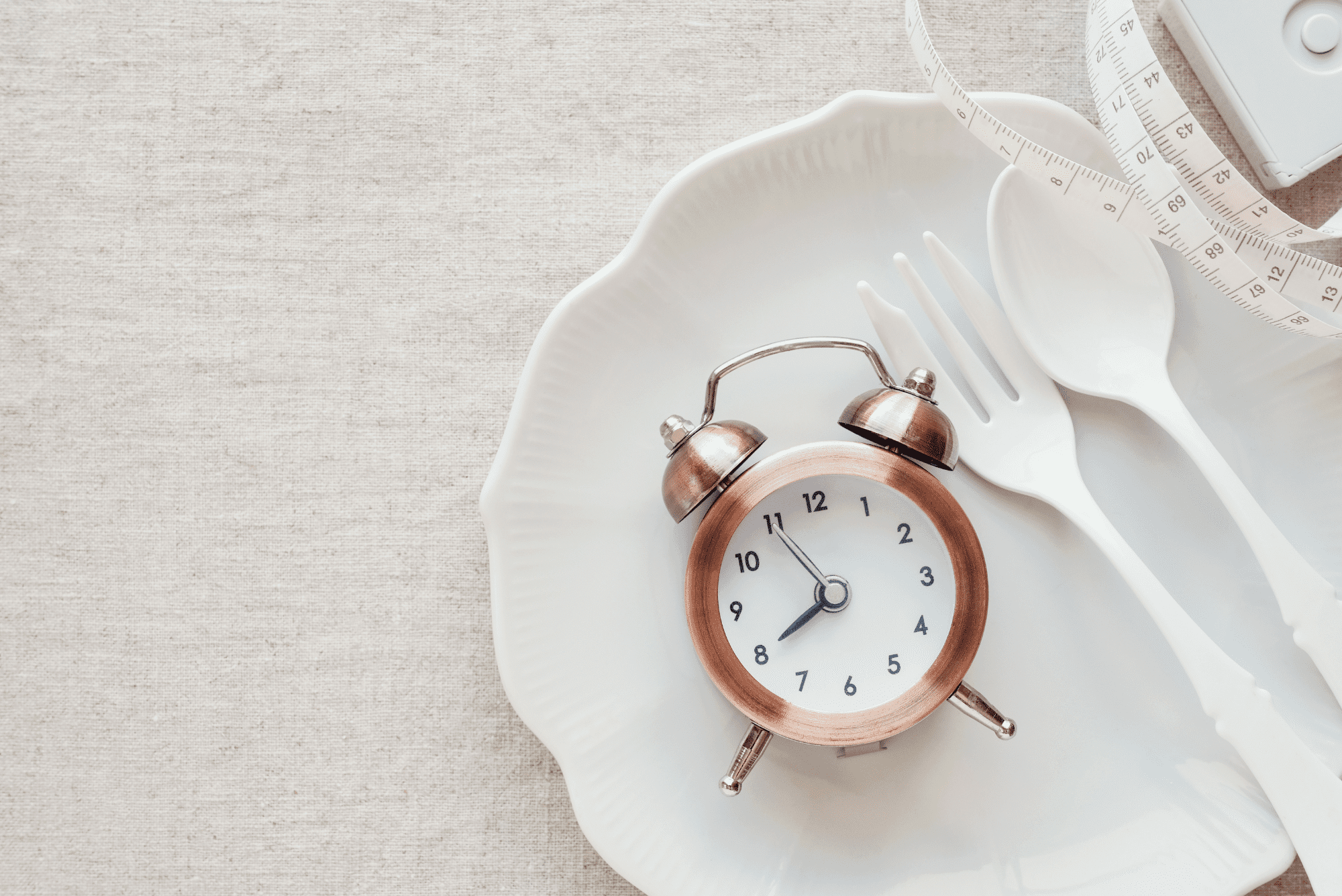 Copper alarm clock on an empty plate with utensils and a measuring tape, symbolizing the fasting‑mimicking diet.