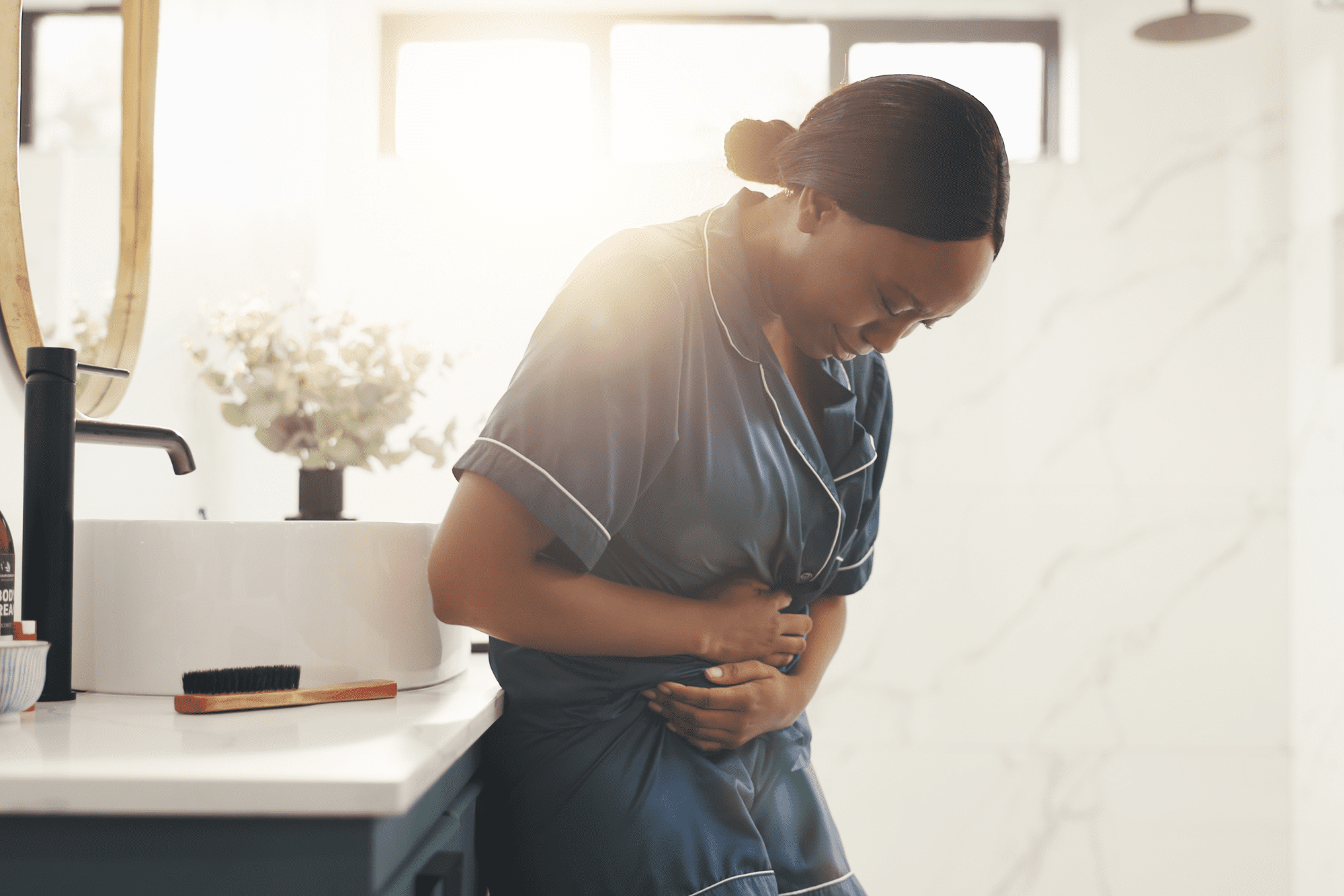 Woman in pajamas holding her stomach in discomfort while leaning against a bathroom counter.