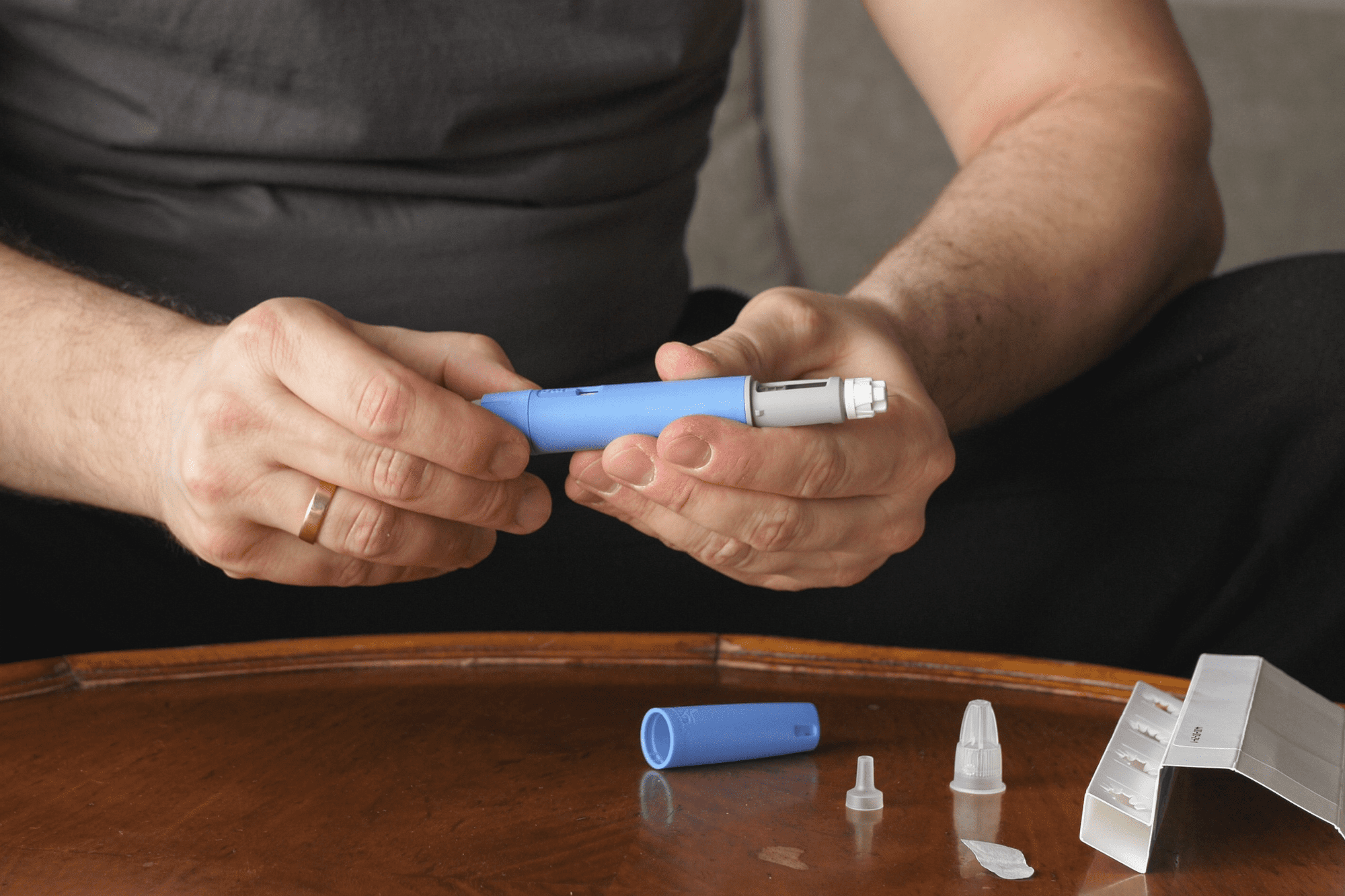 Close-up of hands preparing a blue injectable pen with needle tips and a pill organizer on a wooden side table.